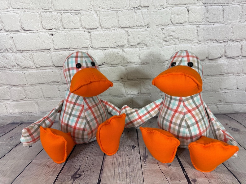 Pu&ograve; includere: Due giocattoli a forma di anatra di peluche con becchi e piedi arancioni. Le anatre hanno corpi a fantasia a quadri rossi, blu e marroni. I giocattoli sono su una superficie di legno davanti a un muro di mattoni bianchi.