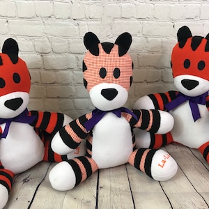 May include: Three plush tiger toys with black and orange stripes. The tigers have white faces with black eyes and noses. The tigers are wearing purple ribbons around their necks. The tigers are sitting on a wooden surface. The text "La-La" is embroidered on the bottom of the tigers.
