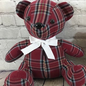 May include: A stuffed teddy bear crafted from a red, black, and grey plaid fabric. The bear features two round, dark brown plastic eyes and a matching dark brown plastic nose. A crisp, white ribbon is tied into a bow around the bear's neck.