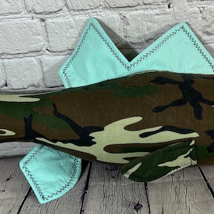 Op de afbeelding: Een pluche vis speelgoed gemaakt van camouflage stof met lichtblauwe vinnen en een zwart knoop oog.