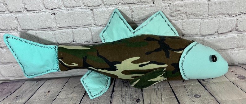 Op de afbeelding: Een pluche vis speelgoed gemaakt van camouflage stof met lichtblauwe vinnen en een zwart knoop oog.