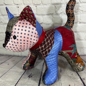 May include: A patchwork cat toy made from different patterned fabrics, including pink with white polka dots, blue with a diamond pattern, and red with a floral design. The cat has a red collar and black eyes.