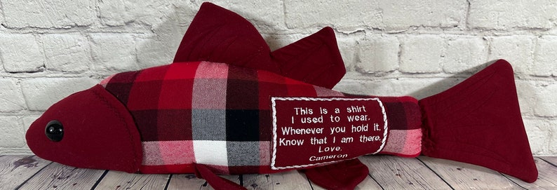 Op de afbeelding: Rood en wit geruit visvormig kussen met een rode viltstaart en een zwart oog. Het kussen heeft een wit geborduurd label met de tekst "This is a shirt I used to wear. Whenever you hold it. Know that I am there. Love, Cameron."