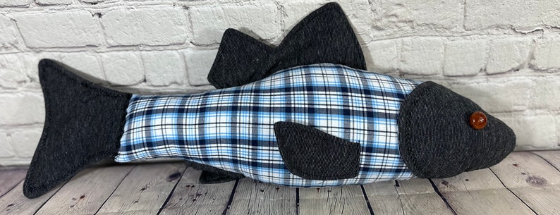 Op de afbeelding: Een grijs en blauw geruit kussen in de vorm van een vis met een bruin knoopje als oog.