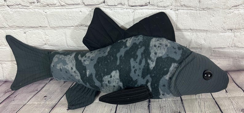 Op de afbeelding: Een grijze en zwarte pluche vis met een camouflagepatroon. De vis heeft een grote staartvin en twee rugvinnen. De vis heeft een knoopoog.