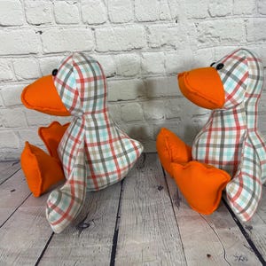 Pu&ograve; includere: Due giocattoli a forma di anatra di peluche con becco e zampe arancioni. I corpi delle anatre sono ricoperti da un motivo a quadri rossi, blu e beige. Sono posizionati su una superficie di legno davanti a un muro di mattoni bianchi.