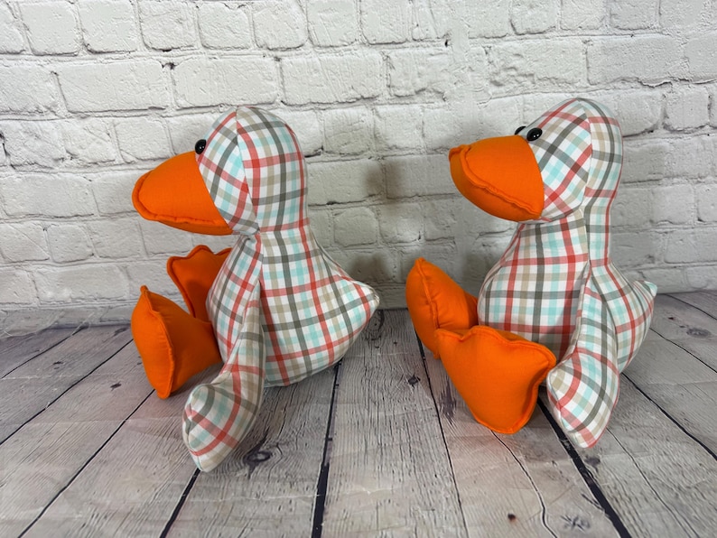 Pu&ograve; includere: Due giocattoli a forma di anatra di peluche con becco e zampe arancioni. I corpi delle anatre sono ricoperti da un motivo a quadri rossi, blu e beige. Sono posizionati su una superficie di legno davanti a un muro di mattoni bianchi.