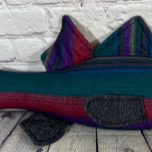 Op de afbeelding: Een pluche vis met een gestreept lichaam in tinten teal, paars en bordeauxrood. De vis heeft een lichtblauwe buik en een zwarte staartvin.