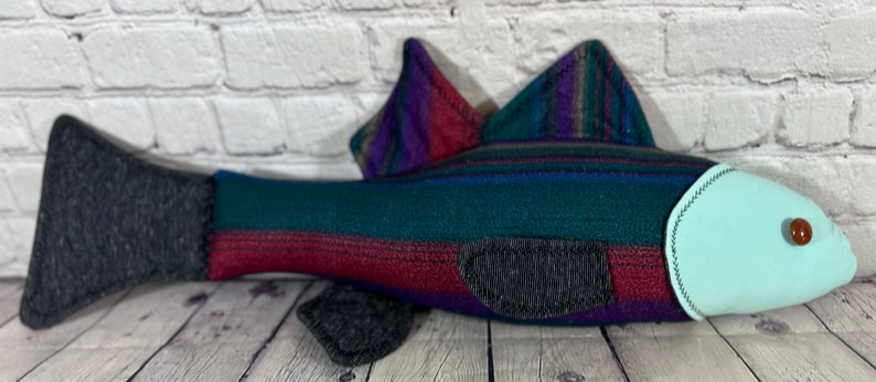 Op de afbeelding: Een pluche vis met een gestreept lichaam in tinten teal, paars en bordeauxrood. De vis heeft een lichtblauwe buik en een zwarte staartvin.