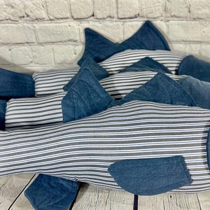 Op de afbeelding: Drie blauwe en witte gestreepte visvormige kussens. De kussens zijn gemaakt van een zachte, pluche stof en hebben een denim-achtige textuur. De kussens zijn in een rij gerangschikt, met het grootste kussen achteraan en het kleinste kussen vooraan.