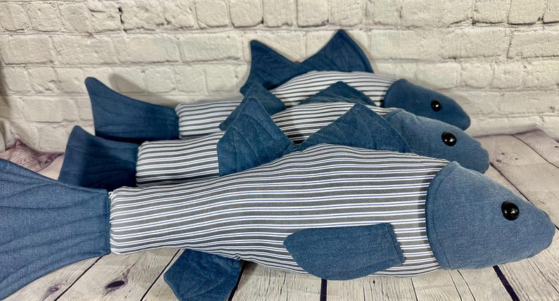 Op de afbeelding: Drie blauwe en witte gestreepte visvormige kussens. De kussens zijn gemaakt van een zachte, pluche stof en hebben een denim-achtige textuur. De kussens zijn in een rij gerangschikt, met het grootste kussen achteraan en het kleinste kussen vooraan.