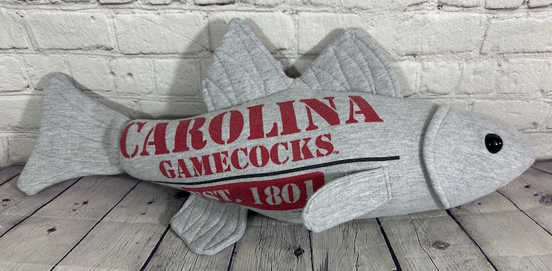 Op de afbeelding: Grijze pluche vis met een rood en wit "Carolina Gamecocks Est. 1801" logo.