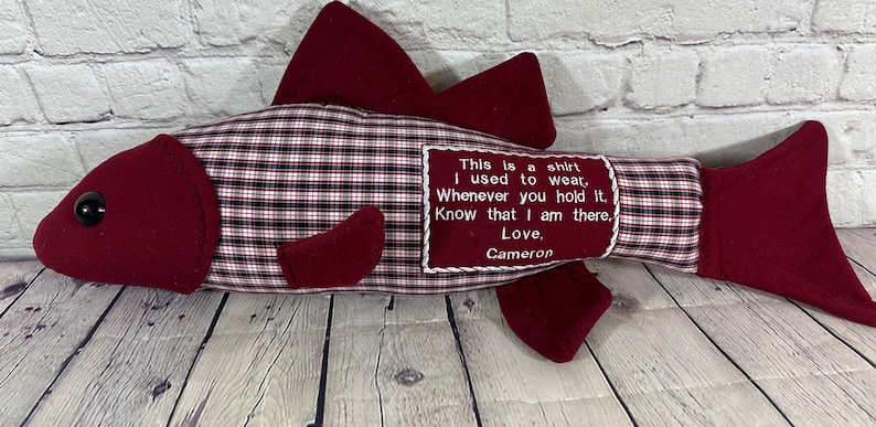 Op de afbeelding: Rode en witte geruite stoffen vis met een rood vilt hoofd en staart. De vis heeft een zwart knoop oog en een geborduurde boodschap met de tekst: "This is a shirt I used to wear, Whenever you hold it, Know that I am there, Love, Cameron."