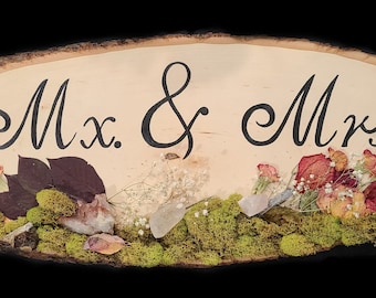 Könnte beinhalten: Ein Holzschild mit dem Text "Mx. & Mrs." in schwarzer Schrift. Das Schild ist mit getrockneten Blumen und Moos verziert.