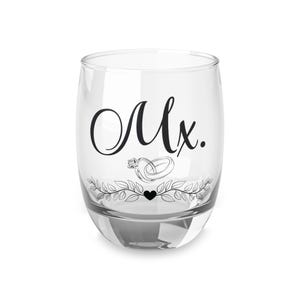 May include: A clear, stemless glass with the word "Mx." in elegant black script. Below the text is a graphic of two intertwined rings, a leafy garland, and a heart. The glass is likely for a wedding or engagement.