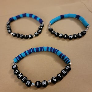 Op de afbeelding: Drie blauwe en zwarte kralenarmbanden met de woorden "STEM Girls Rule", "Feminist" en "Women in STEM" erop.