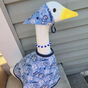 May include: A decorative goose wearing a light blue outfit with a matching hat and a blue beaded necklace. The outfit and hat feature the text "North Carolina" and various graphics in white. The goose has a yellow beak.
