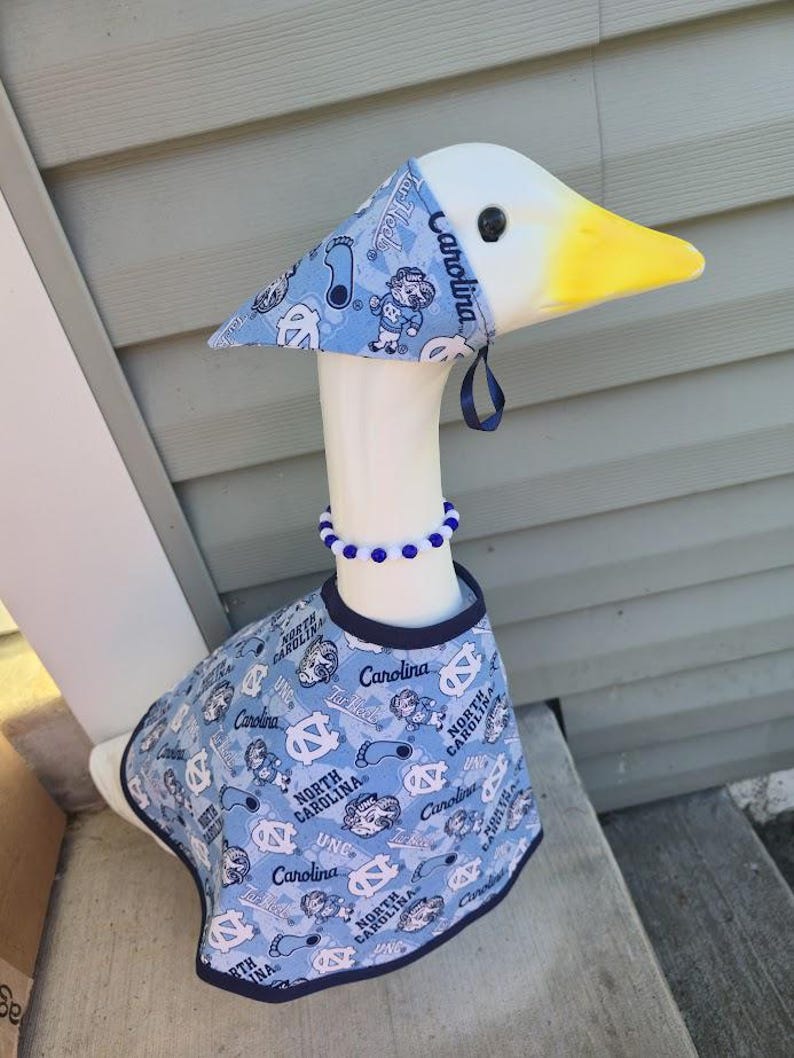 May include: A decorative goose wearing a light blue outfit with a matching hat and a blue beaded necklace. The outfit and hat feature the text "North Carolina" and various graphics in white. The goose has a yellow beak.