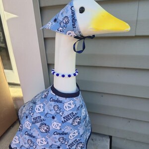 May include: A decorative goose figurine wearing a light blue outfit with a matching hat and beaded necklace. The outfit features the text "Carolina" and "North Carolina" with Tar Heel logos.