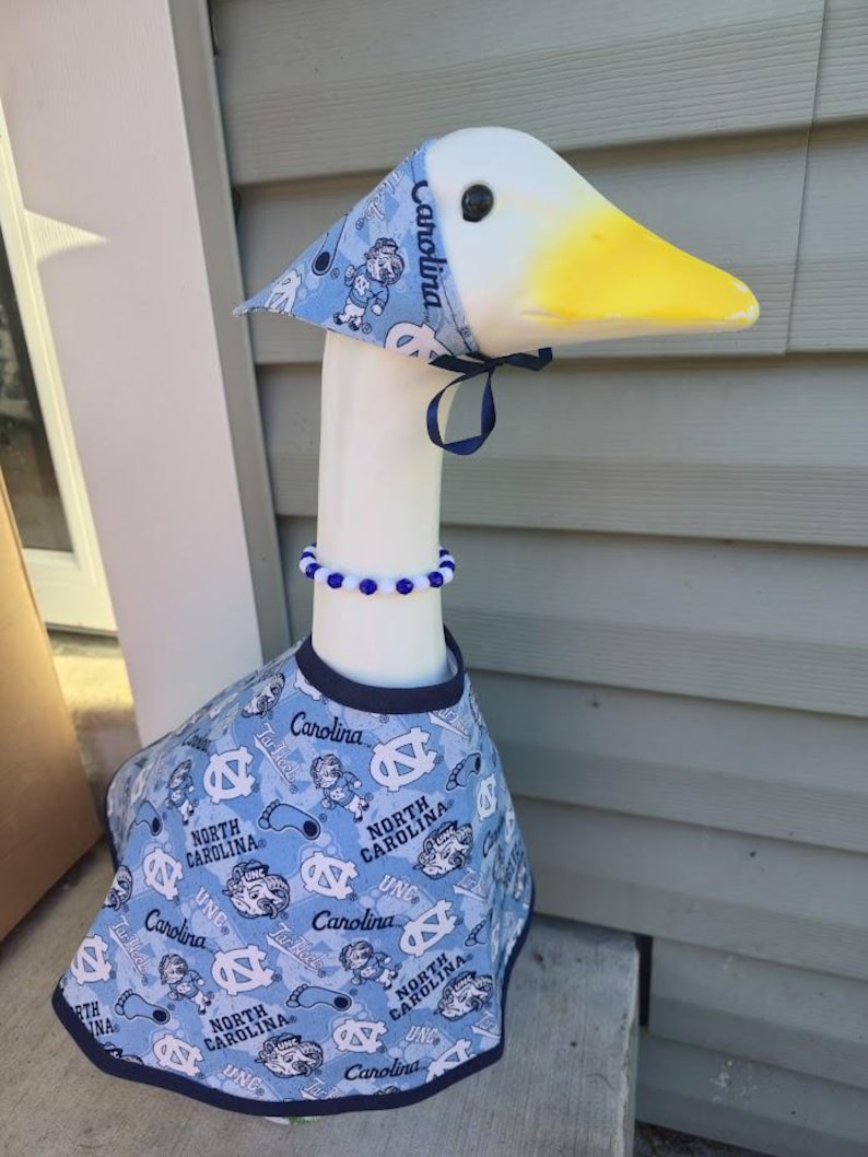 May include: A decorative goose figurine wearing a light blue outfit with a matching hat and beaded necklace. The outfit features the text "Carolina" and "North Carolina" with Tar Heel logos.