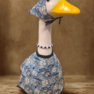 May include: A decorative goose figurine wearing a light blue outfit with the words "North Carolina" and "UNC" printed on it. The goose has a matching hat and a beaded necklace. The goose has a white head, a yellow beak, and a black eye.