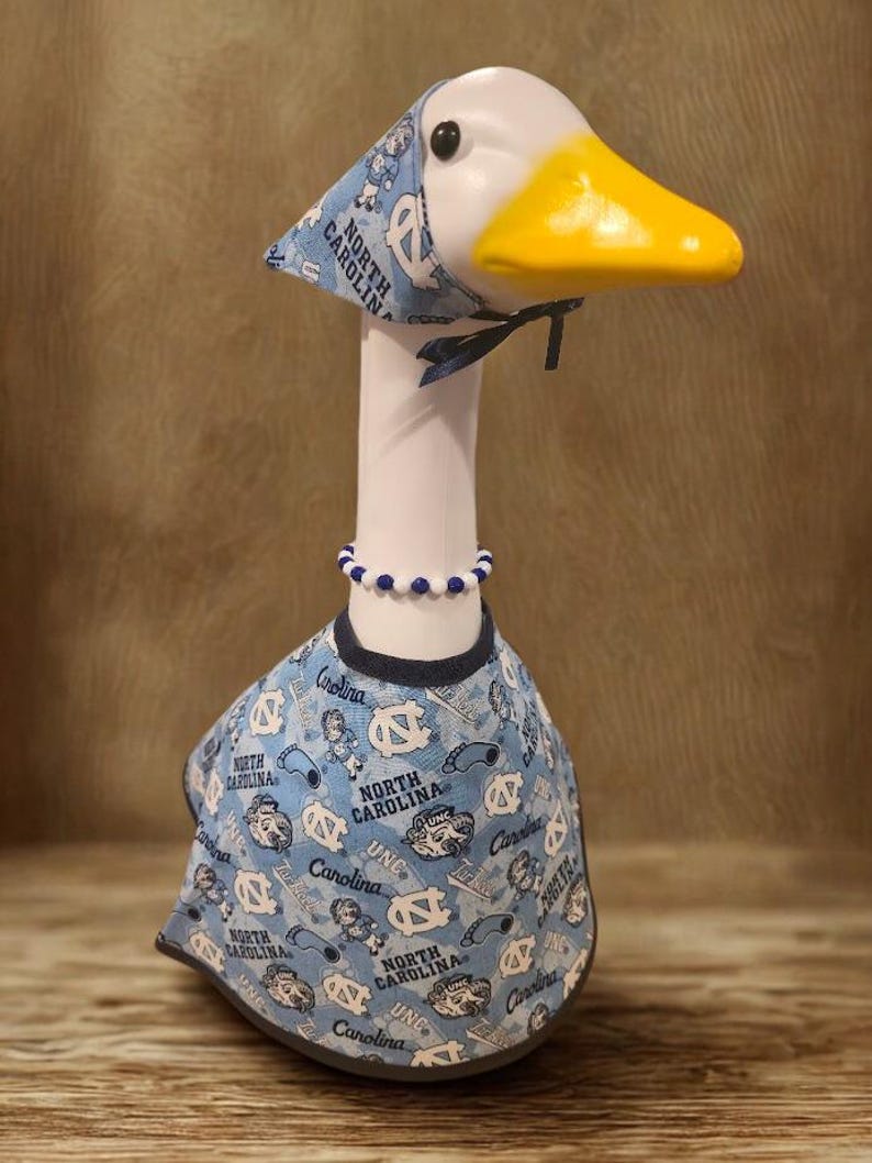 May include: A decorative goose figurine wearing a light blue outfit with the words "North Carolina" and "UNC" printed on it. The goose has a matching hat and a beaded necklace. The goose has a white head, a yellow beak, and a black eye.