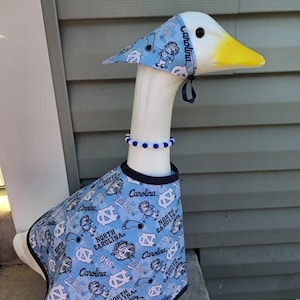 May include: A decorative goose figurine wearing a light blue outfit with a matching hat and beaded necklace. The outfit features the text "Carolina" and "North Carolina" with various graphics. The goose has a white body, yellow beak, and black eyes.