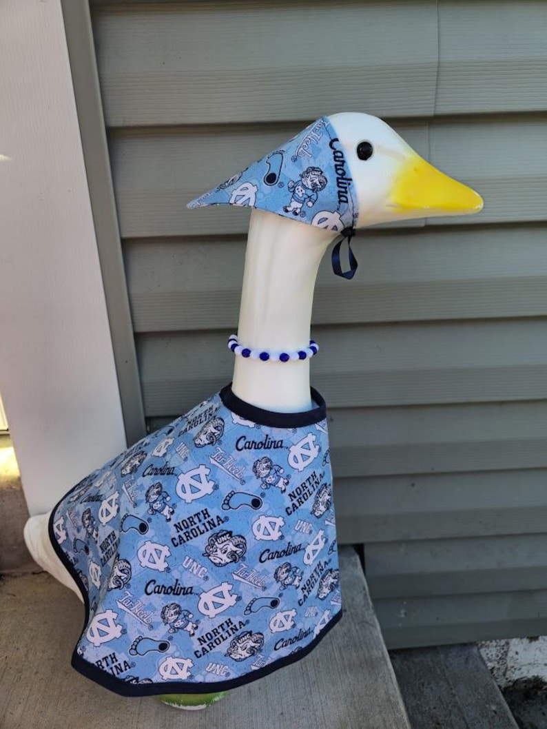 May include: A decorative goose figurine wearing a light blue outfit with a matching hat and beaded necklace. The outfit features the text "Carolina" and "North Carolina" with various graphics. The goose has a white body, yellow beak, and black eyes.
