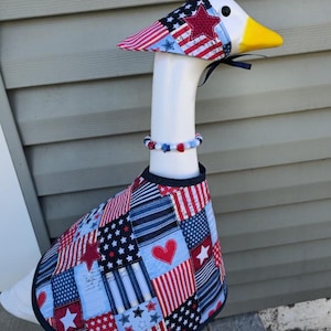 May include: A decorative goose figurine wearing a patriotic outfit. The outfit includes a hat, collar, and vest, all adorned with red, white, and blue patterns featuring stars and stripes. The goose has a yellow beak and a white neck.