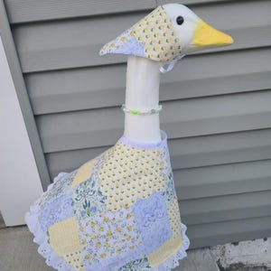 May include: A whimsical goose statue with a white neck and head, featuring a yellow beak and a patterned bonnet. The goose wears a patchwork dress in yellow, blue, and white, trimmed with white lace. A beaded necklace adorns its neck.
