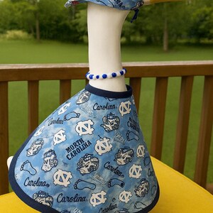 May include: A ceramic goose figurine wearing a blue and white outfit with the text "North Carolina" and "Carolina". The goose has a white body, yellow beak, and a blue and white beaded necklace.