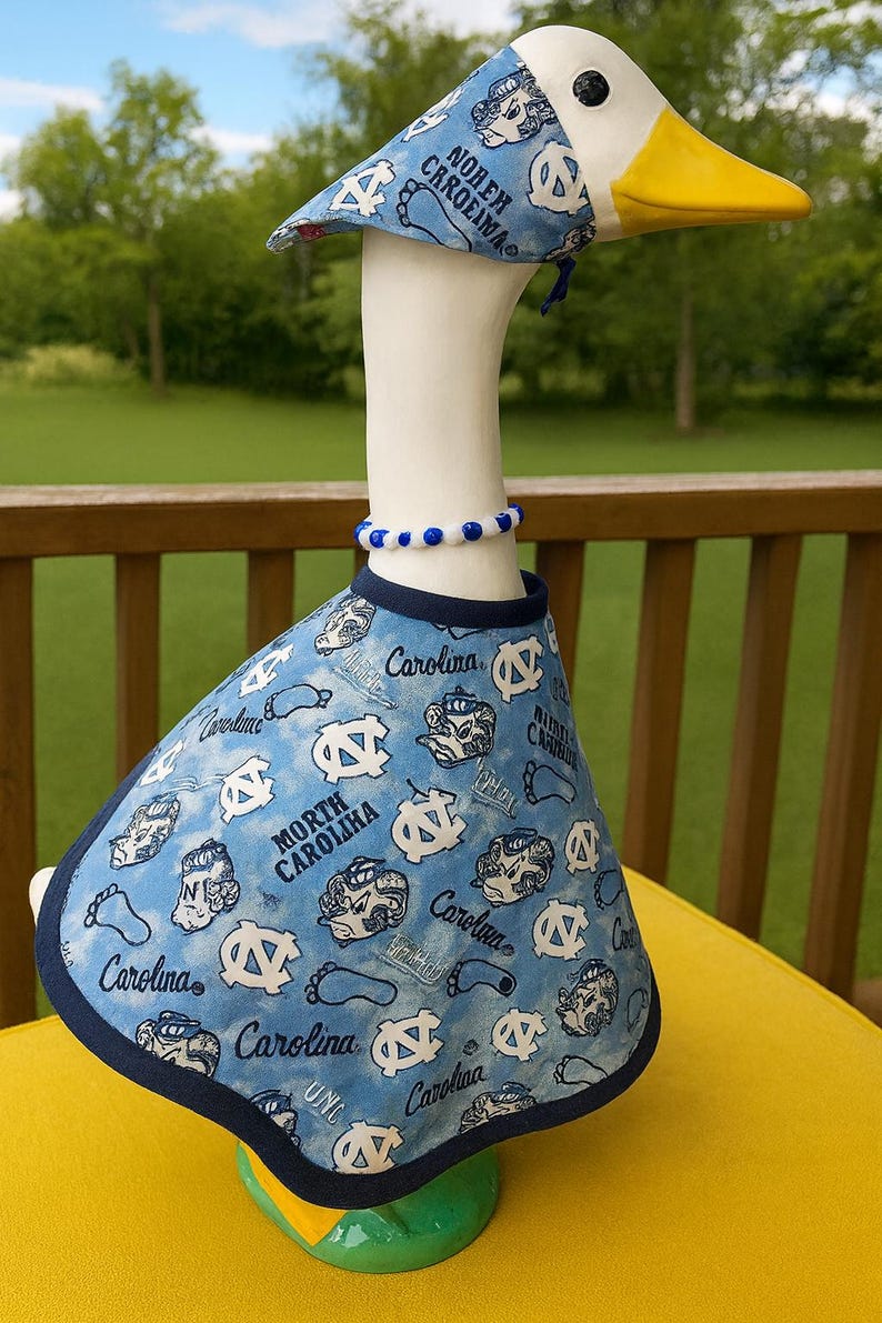 May include: A ceramic goose figurine wearing a blue and white outfit with the text "North Carolina" and "Carolina". The goose has a white body, yellow beak, and a blue and white beaded necklace.
