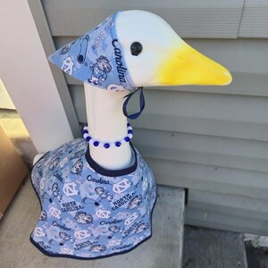 May include: A decorative goose figurine with a white head and neck, a yellow beak, and a blue and white patterned outfit. The outfit includes a hat, bib, and beaded necklace. The pattern features the text "Carolina" and cartoon figures.