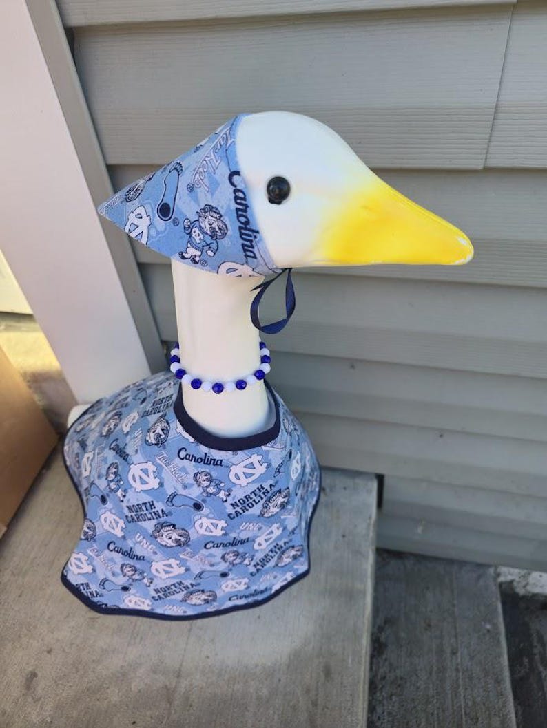 May include: A decorative goose figurine with a white head and neck, a yellow beak, and a blue and white patterned outfit. The outfit includes a hat, bib, and beaded necklace. The pattern features the text "Carolina" and cartoon figures.