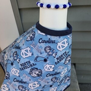 May include: A light blue pet shirt with a navy blue trim, patterned with white logos, text, and cartoon figures. A matching blue and white beaded bracelet is worn above the shirt. The shirt features the text "Carolina" and "North Carolina".