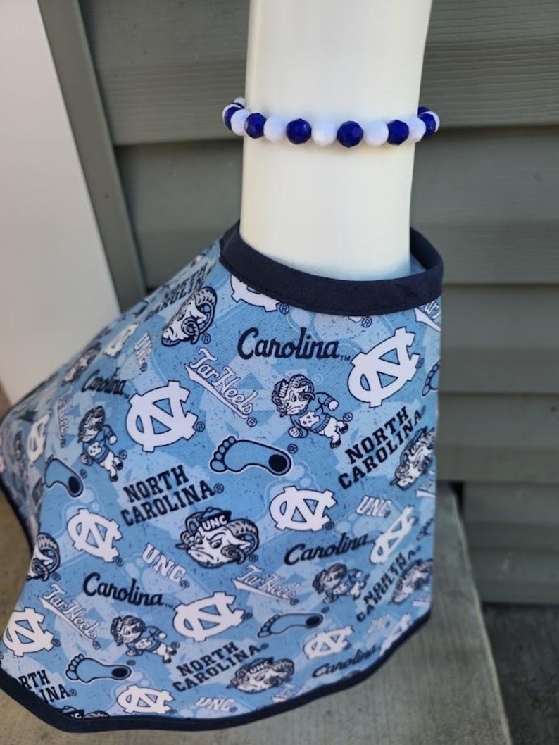 May include: A light blue pet shirt with a navy blue trim, patterned with white logos, text, and cartoon figures. A matching blue and white beaded bracelet is worn above the shirt. The shirt features the text "Carolina" and "North Carolina".