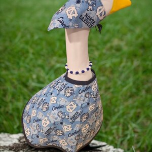 May include: A decorative goose figurine wearing a light blue outfit with a pattern of logos and text that reads "North Carolina" and "Carolina". The goose has a white head, a yellow beak, and a beaded necklace.