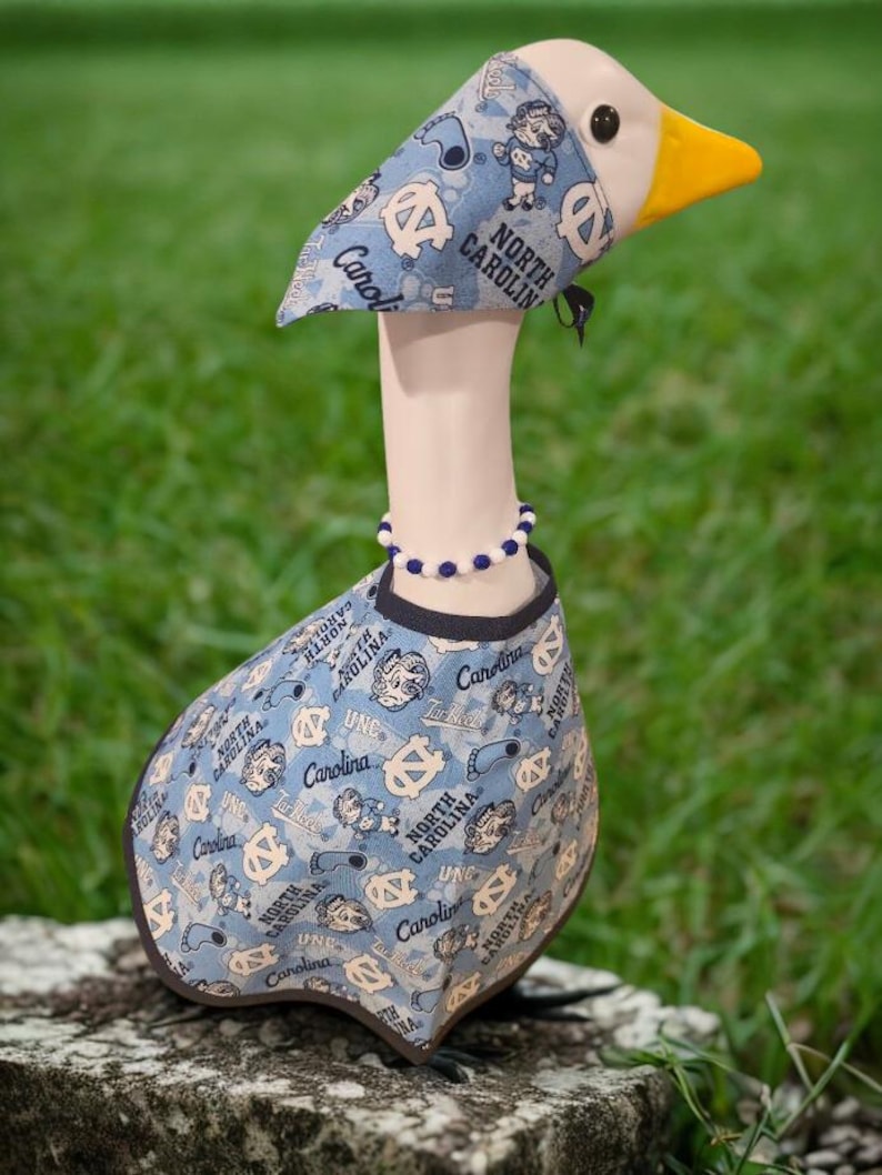 May include: A decorative goose figurine wearing a light blue outfit with a pattern of logos and text that reads "North Carolina" and "Carolina". The goose has a white head, a yellow beak, and a beaded necklace.