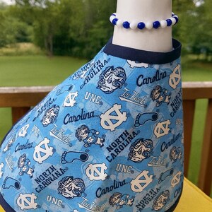 May include: A light blue pet shirt with a navy blue trim, patterned with white logos and text including "UNC", "Tar Heels", and "Carolina". A blue and white beaded bracelet is worn on the neck.