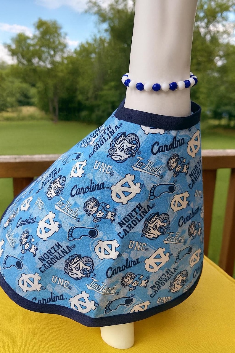 May include: A light blue pet shirt with a navy blue trim, patterned with white logos and text including "UNC", "Tar Heels", and "Carolina". A blue and white beaded bracelet is worn on the neck.