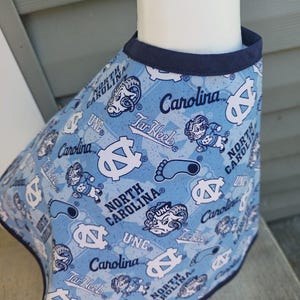 May include: Light blue pet bandana featuring a repeating pattern of UNC logos, Tar Heels text, and the words "North Carolina" in white. The bandana has a navy blue trim and is displayed on a white form. A blue and white beaded bracelet is visible.