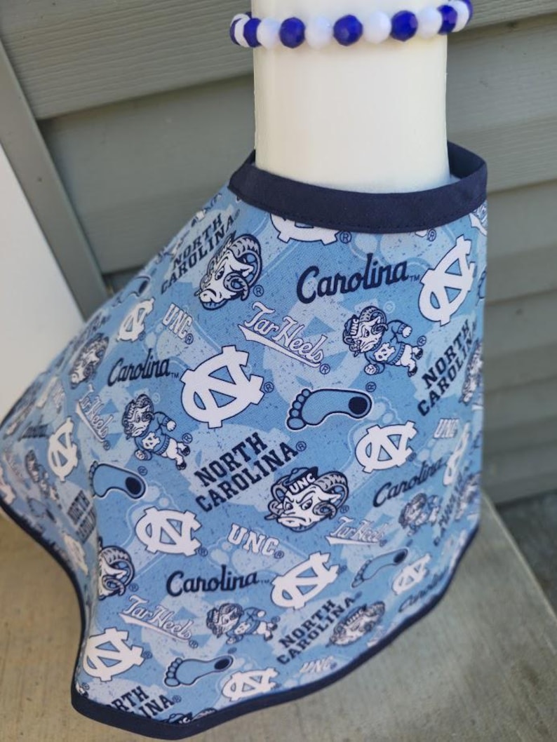 May include: Light blue pet bandana featuring a repeating pattern of UNC logos, Tar Heels text, and the words "North Carolina" in white. The bandana has a navy blue trim and is displayed on a white form. A blue and white beaded bracelet is visible.