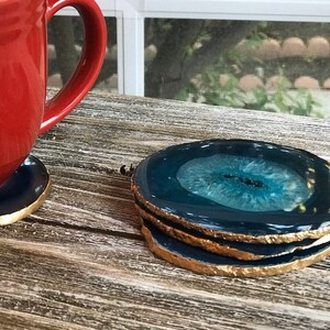 May include: A stack of three agate coasters with a dark blue and teal center and gold-painted edges. A red mug sits on a matching coaster. The coasters are on a weathered wooden surface.
