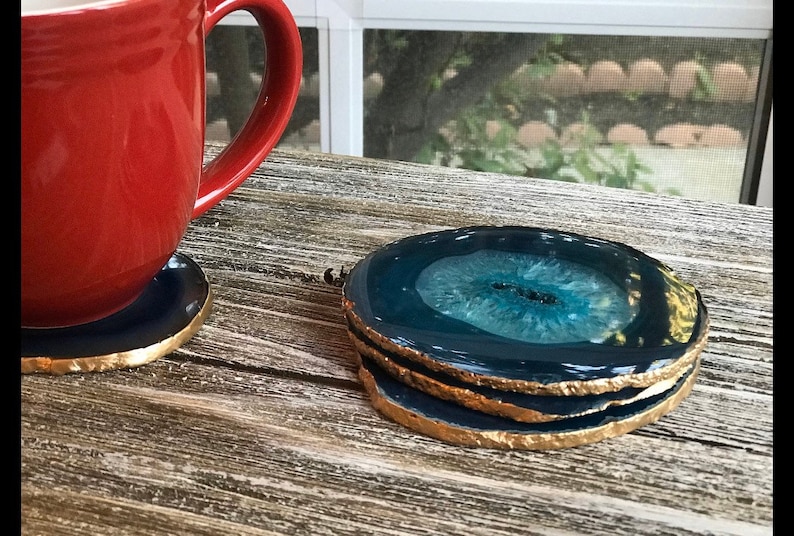 May include: A stack of three agate coasters with a dark blue and teal center and gold-painted edges. A red mug sits on a matching coaster. The coasters are on a weathered wooden surface.