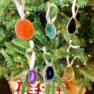 May include: Six agate slice ornaments with silver ribbon hanging on a green Christmas tree. The ornaments are in various colors, including orange, green, black, purple, blue, and yellow.