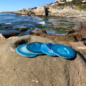 May include: Four blue agate coasters with white and gray banding, arranged on a rocky beach with the ocean in the background.