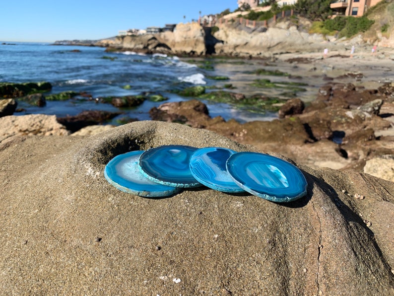 May include: Four blue agate coasters with white and gray banding, arranged on a rocky beach with the ocean in the background.