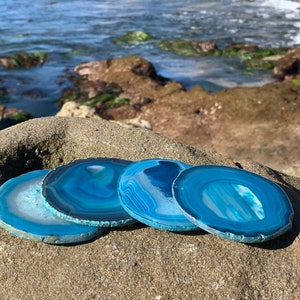 May include: Four blue agate coasters with white and gray banding. The coasters are arranged in a semi-circle on a rocky surface.