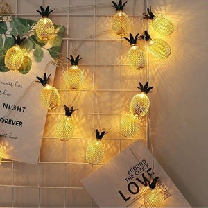 May include: String of lights with pineapple-shaped bulbs. The lights are strung on a wire and are lit up. The pineapples are gold and have a black top. The lights are hanging on a white grid wall. The text "Around the clock love" is visible on a white card.