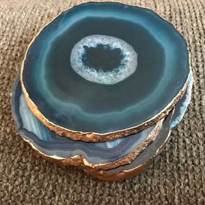 May include: A stack of four blue agate coasters with gold-painted edges. The coasters have a circular shape with a white and blue center, and are stacked on top of each other.
